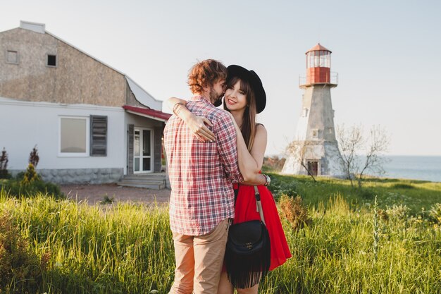 Abrazando a la joven pareja elegante enamorada en el campo, estilo bohemio indie hipster, vacaciones de fin de semana, traje de verano, vestido rojo, hierba verde, tomados de la mano