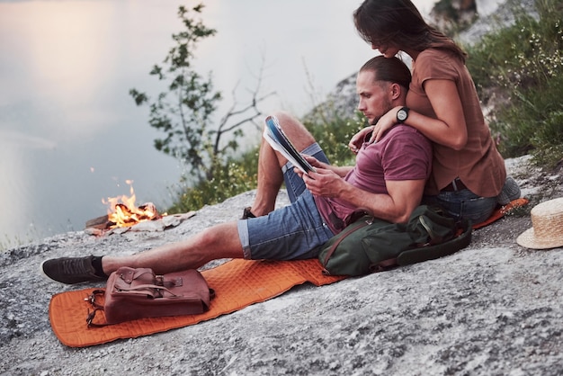 Abrazando pareja con mochila sentado cerca del fuego en la cima de la montaña disfrutando de la vista de la costa de un río o lago.