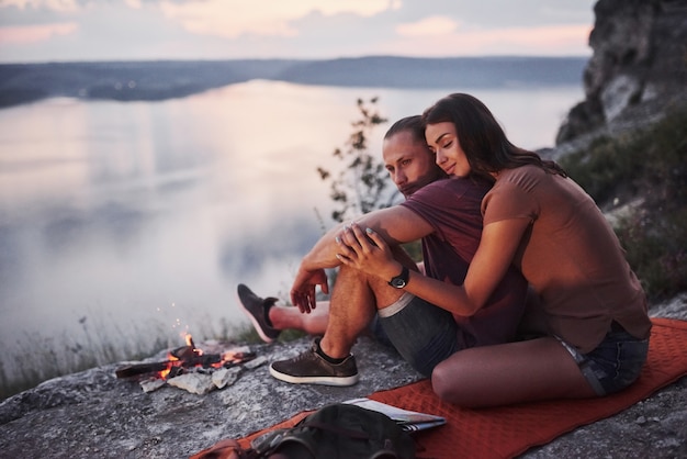 Abrazando pareja con mochila sentado cerca del fuego en la cima de la montaña disfrutando de la vista de la costa de un río o lago.
