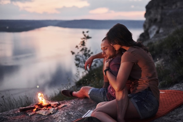 Abrazando pareja con mochila sentado cerca del fuego en la cima de la montaña disfrutando de la vista de la costa de un río o lago.