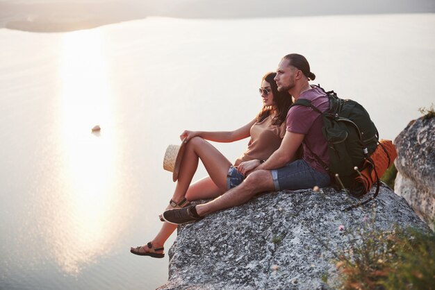 Abrazando pareja con mochila sentado en la cima de la montaña de roca disfrutando de la vista de la costa de un río o lago.