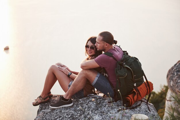 Abrazando pareja con mochila sentado en la cima de la montaña de roca disfrutando de la vista de la costa de un río o lago.