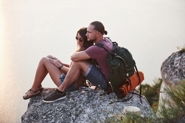 Abrazando pareja con mochila sentado en la cima de la montaña de roca disfrutando de la vista de la costa de un río o lago.