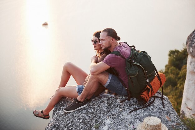 Abrazando pareja con mochila sentado en la cima de la montaña de roca disfrutando de la vista de la costa de un río o lago.