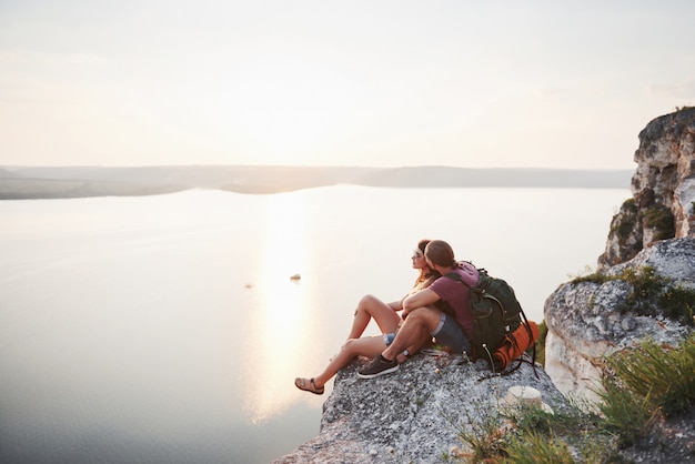 Abrazando pareja con mochila sentado en la cima de la montaña de roca disfrutando de la vista de la costa de un río o lago.