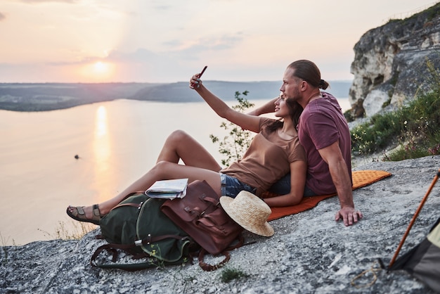 Abrazando pareja con mochila sentado en la cima de la montaña de roca disfrutando de la vista de la costa de un río o lago.