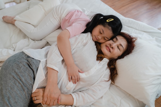 La abuela asiática duerme en casa. Los chinos mayores, abuela feliz se relajan con la muchacha joven de la nieta que besa la mejilla para despertar acostado en cama en dormitorio en casa en el concepto de la noche.