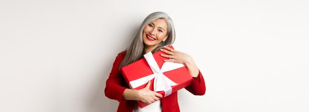 Abuela asiática feliz abrazando una caja de regalo roja y sonriendo agradecida agradeciendo el regalo de pie