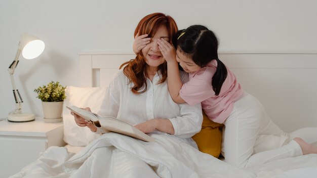 Abuela asiática relajarse en casa. Los chinos mayores, la abuela feliz se relajan con la muchacha joven de la nieta que disfruta cerca de su sorpresa de los ojos que juega junto que miente en cama en dormitorio en casa en el concepto de la noche.