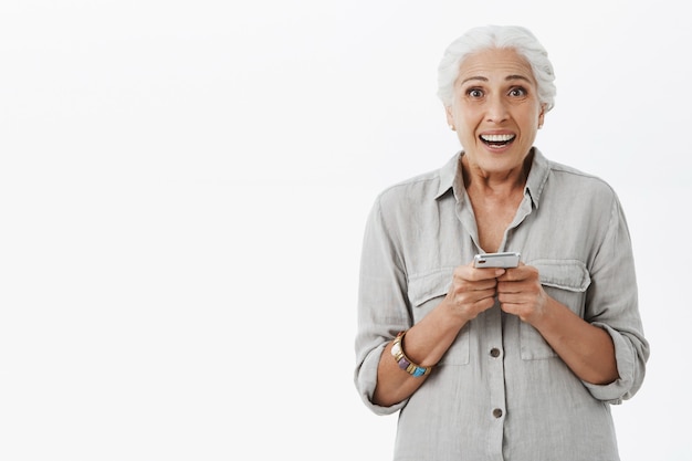 Abuela emocionada y sorprendida sosteniendo el teléfono móvil y sonriendo feliz