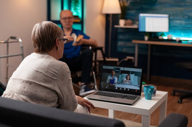 Abuela llamando al médico en la clínica de la sala del hospital para verificar el diagnóstico de atención médica en la conferencia de videoconferencia. Mujer hablando con el médico sobre el tratamiento de su sobrina mientras el hombre se sienta en silla de ruedas