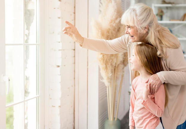 Abuela con niña en casa mirando por la ventana