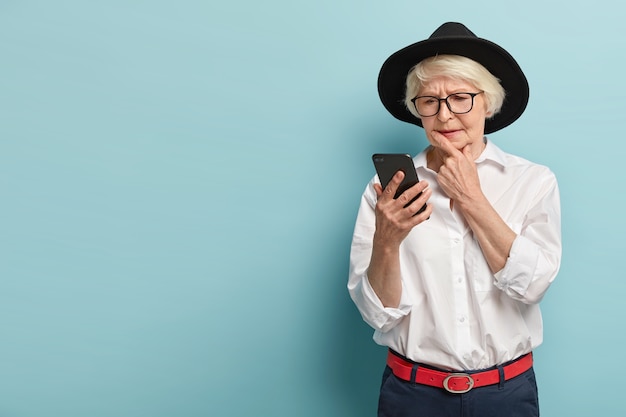 La abuela seria concentrada usa tocados de moda, atuendo, enfocada en el teléfono inteligente, lee atentamente noticias en línea, modelos sobre una pared azul con espacio libre, revisa el buzón de correo electrónico. Pensionista moderno