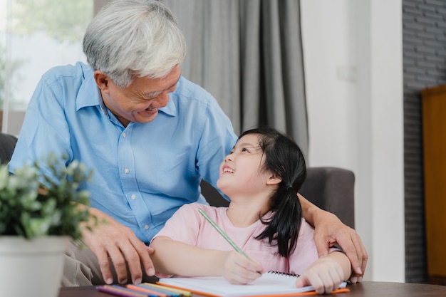 El abuelo asiático enseña a la nieta a dibujar y hacer los deberes en casa. Los chinos mayores, abuelo feliz se relajan con el concepto de la chica joven que miente en el sofá en sala de estar en casa.