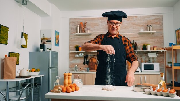 El abuelo espolvoreando sobre la mesa tamizó la harina en la cocina moderna. Anciano panadero senior con bonete y tamizado uniforme, tamizado, esparcimiento de ingredientes rew en masa, horneado de pizza y pan casero.