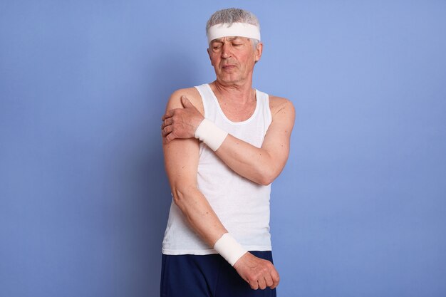 Foto gratuita abuelo jubilado frustrado que tiene un fuerte dolor en la nuca, vestido con camiseta blanca sin mangas, diadema, pretina posando aislada.