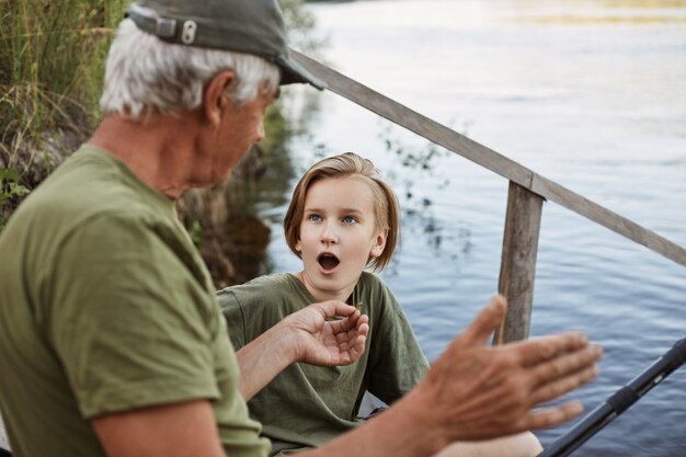 Abuelo y nieto pescando en el muelle del río, un hombre mayor mostrando el tamaño del pez que pescó la última vez, un niño posando con la boca abierta, sorprendido.