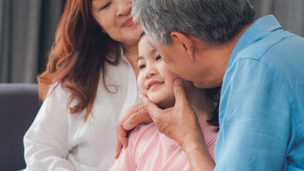 Los abuelos asiáticos besan la mejilla de la nieta en casa. Los chinos mayores, la vieja generación, el abuelo y la abuela que usan el tiempo familiar se relajan con el niño de la chica joven que miente en el sofá en concepto de la sala de estar.