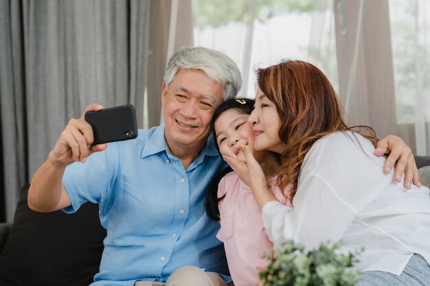 Abuelos asiáticos selfie con nieta en casa. Los chinos mayores, el abuelo y la abuela felices pasan el tiempo en familia relajándose usando el teléfono móvil con el niño de la niña acostado en el sofá en concepto de sala de estar.