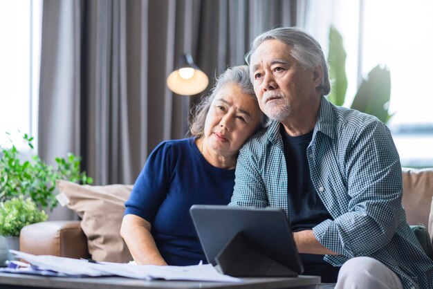 Los abuelos asiáticos se sientan juntos en el sofá con el estrés, la sensación de infelicidad, la deuda del hogar, el problema financiero de la familia, la pareja de ancianos se ve desesperada en el sofá de la sala de estar.