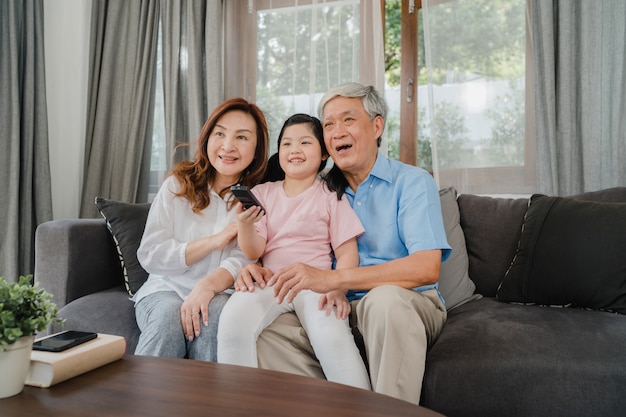 Los abuelos asiáticos ven la televisión con su nieta en casa. El chino mayor, el abuelo y la abuela felices usando tiempo de la familia se relajan con el niño de la chica joven que miente en el sofá en concepto de la sala de estar.