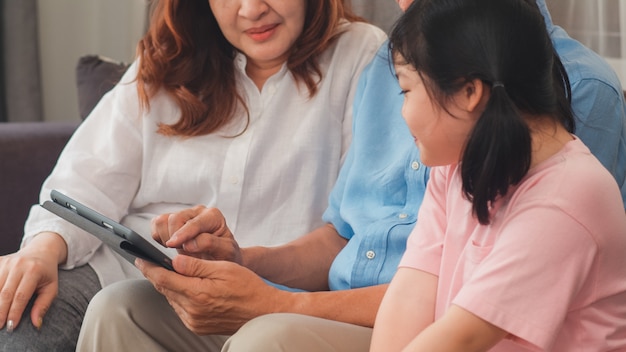 Abuelos y nieta asiáticos que usan la tableta en casa. Senior chino, abuelo y abuela feliz pasar tiempo familiar relajarse con una niña revisando las redes sociales, acostado en el sofá en concepto de sala de estar