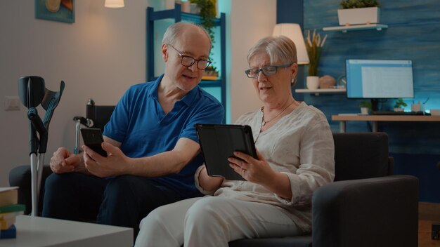 Abuelos que usan teléfonos inteligentes y tabletas digitales en casa, disfrutan de la tecnología en el tiempo libre. Hombre con discapacidad mirando la pantalla del teléfono móvil y comenzando una conversación con su esposa.