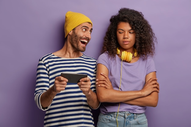 Aburrida, disgustada, insatisfecha, adolescente afroamericana mantiene las manos cruzadas, mira cómo un amigo juega videojuegos en un teléfono inteligente moderno, obsesionado con la nueva aplicación.