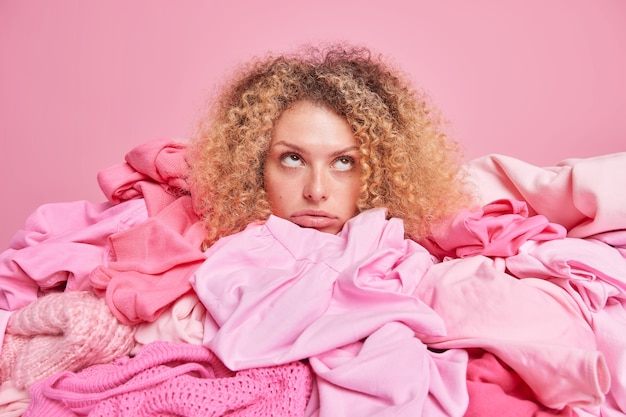 Aburrida mujer joven triste con cabello rizado y tupido posa alrededor de montones de ropa enfocada arriba aislada sobre pared rosa. Armario femenino desordenado. Reciclaje de tejidos y concepto de reutilización textil.