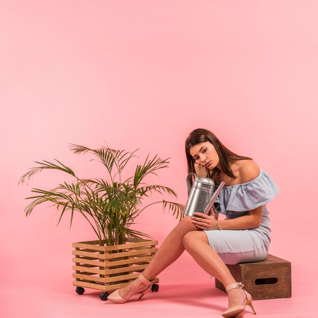 Aburrida mujer sentada con regadera cerca de la planta