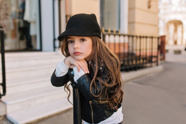 Aburrida niña encantadora con lindo sombrero negro esperando a la madre afuera frente al salón de belleza.