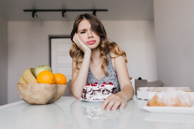 Aburrida señorita sentada a la mesa con pastel y frutas. Chica bastante blanca posando durante el desayuno.