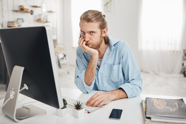 Aburrido empleado de oficina caucásico barbudo con una mirada desesperada que se acerca a la fecha límite, pero no puede terminar el informe a tiempo. Hombre empleado sentado frente a la computadora en la luz, escribiendo informe.