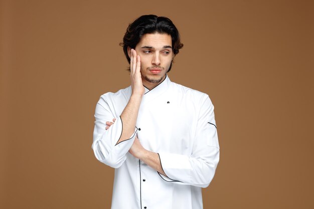 Aburrido joven chef masculino vistiendo uniforme manteniendo la mano en la cara y otra mano en el brazo mirando al lado aislado sobre fondo marrón