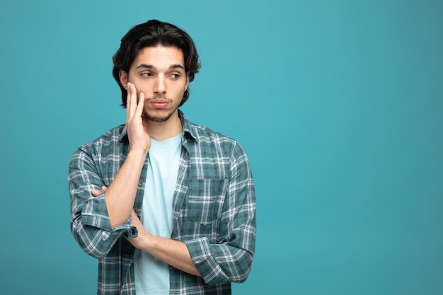 Aburrido joven guapo manteniendo la mano en el brazo y en la cara mirando al lado aislado en fondo azul con espacio de copia