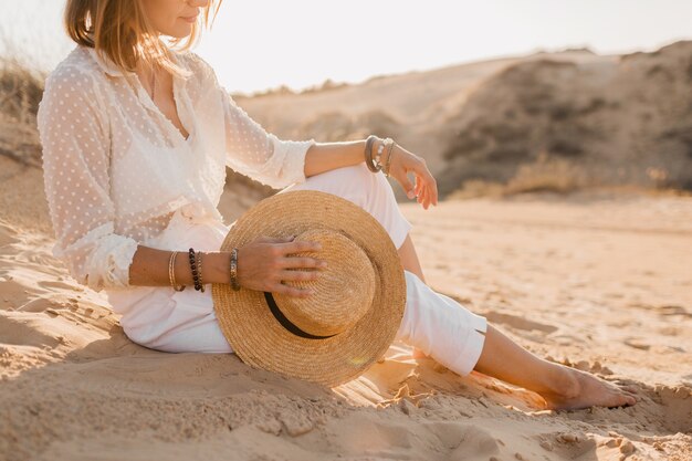 Accesorios de moda de primer plano de mujer hermosa con estilo en la playa del desierto en traje blanco con sombrero de paja en la puesta del sol