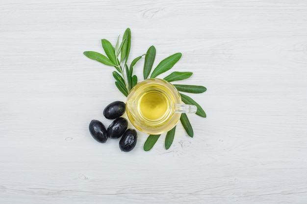 Aceitunas negras y aceite de oliva en un frasco de vidrio con vista superior de hojas de olivo sobre tablón de madera blanca
