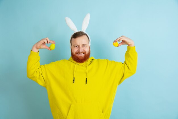 Se acerca la búsqueda de huevos. Hombre caucásico como un conejito de Pascua con ropa casual brillante sobre fondo azul de estudio.