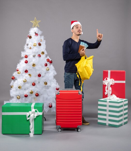 Aclamando a joven con gorro de Papá Noel y maleta roja comprobando su mochila amarilla alrededor de diferentes regalos en gris