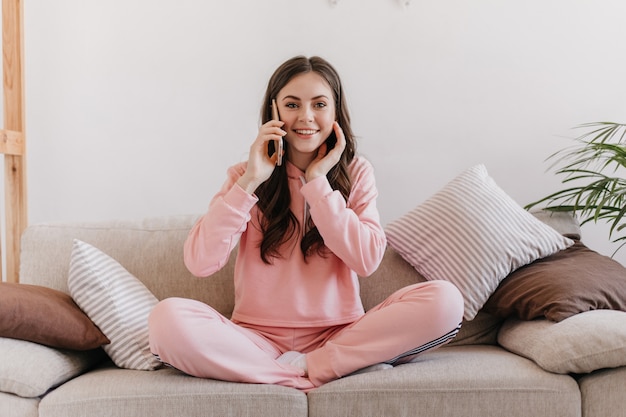 En el acogedor apartamento hermosa mujer con maquillaje suave en su rostro y dulce sonrisa habla en su teléfono