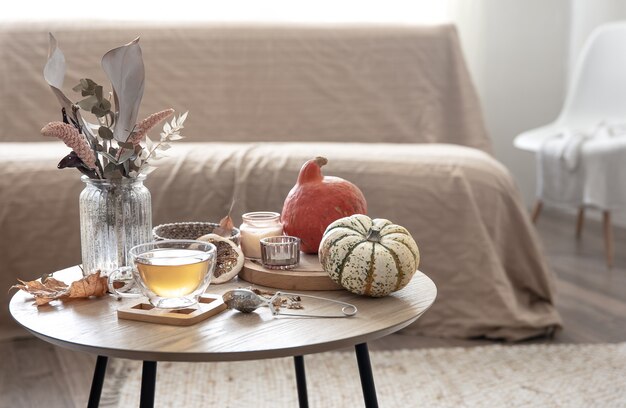 Acogedor bodegón con una taza de té, calabazas, velas y detalles de decoración otoñal sobre una mesa sobre un fondo borroso de la habitación.