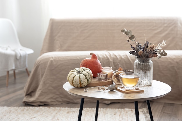 Acogedor bodegón con una taza de té, calabazas, velas y detalles de decoración otoñal sobre una mesa sobre un fondo borroso de la habitación.