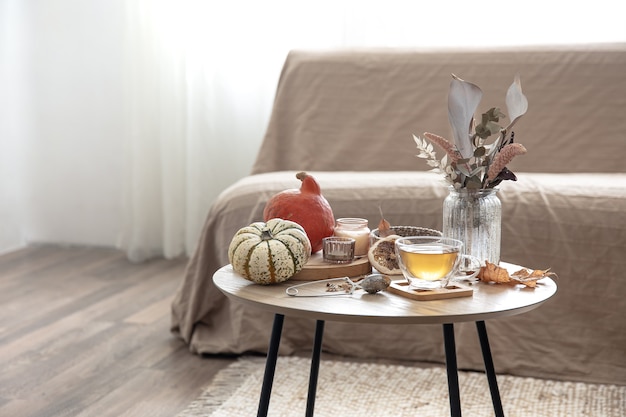 Acogedor bodegón con una taza de té, calabazas, velas y detalles de decoración otoñal sobre una mesa sobre un fondo borroso de la habitación.