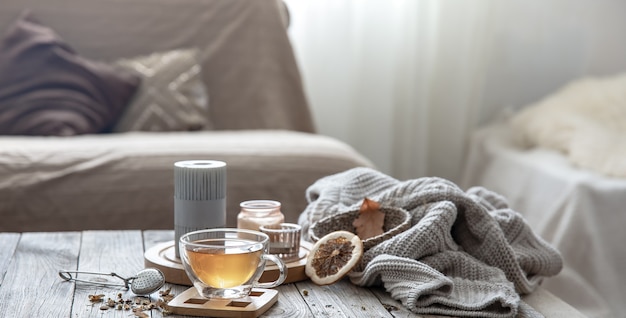 Acogedora composición de hogar otoñal con una taza de té, velas y un elemento de punto sobre un fondo borroso del interior de la habitación.