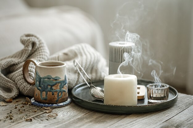 Acogedora composición con una taza de cerámica, velas y un elemento de punto sobre un fondo difuminado.