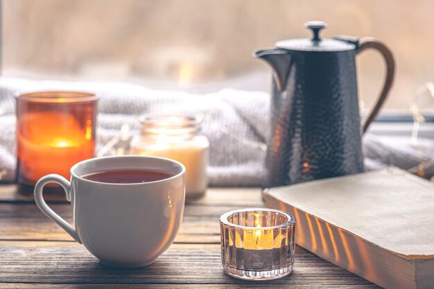 Acogedora composición con una taza de té, un libro y velas junto a la ventana