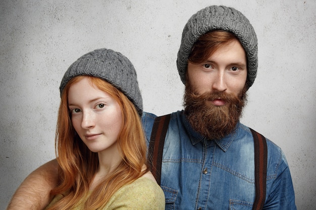 Acogedora foto de dos modelos caucásicos con sombreros de punto gris posando juntos