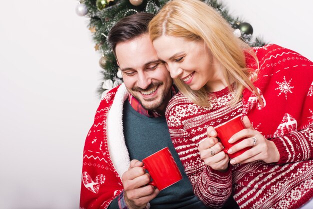 Acogedora foto de una pareja feliz bebiendo té caliente en el fondo del árbol de Navidad