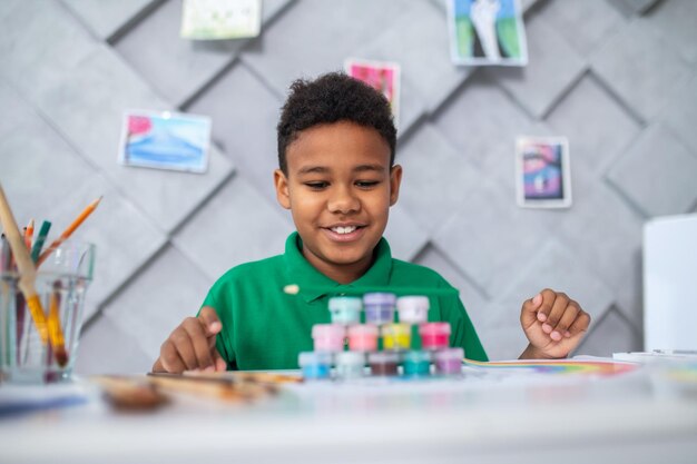 Actividad interesante. Sonriente niño negro en edad escolar sentado en la mesa mirando tubos de pintura apilados en pirámide en una habitación luminosa con luz del día