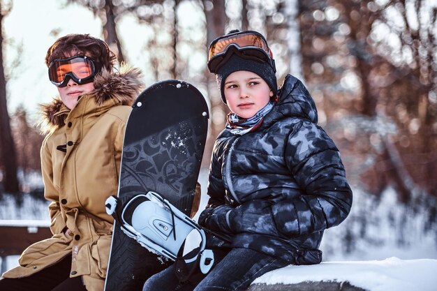 Actividades al aire libre durante las vacaciones de invierno. Dos hermanos adolescentes vestidos con ropa de abrigo sentados en un banco con una tabla de snowboard en un parque forestal